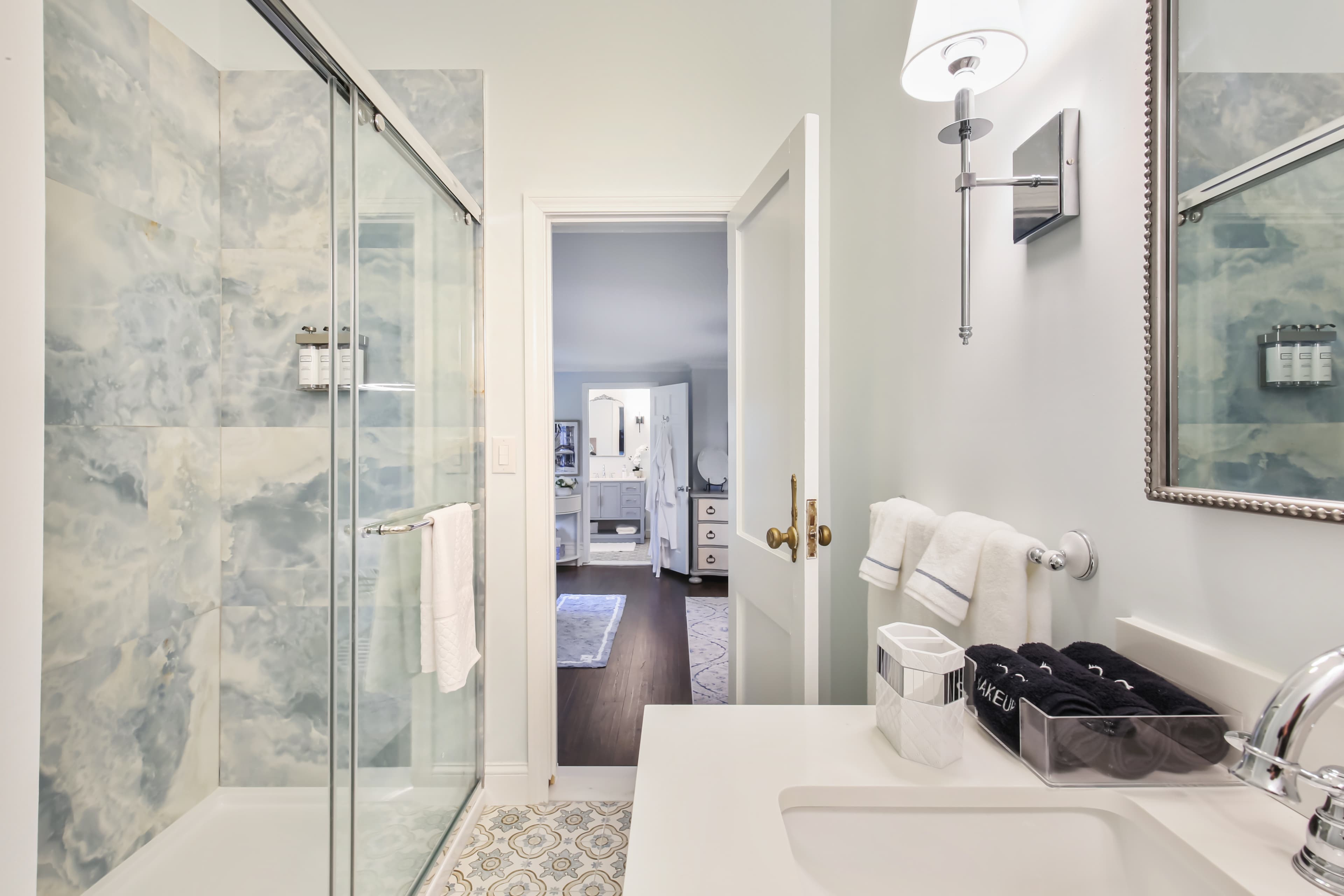 A modern bathroom featuring a glass shower, a sink with toiletries, and an open door leading to a spacious room beyond.
