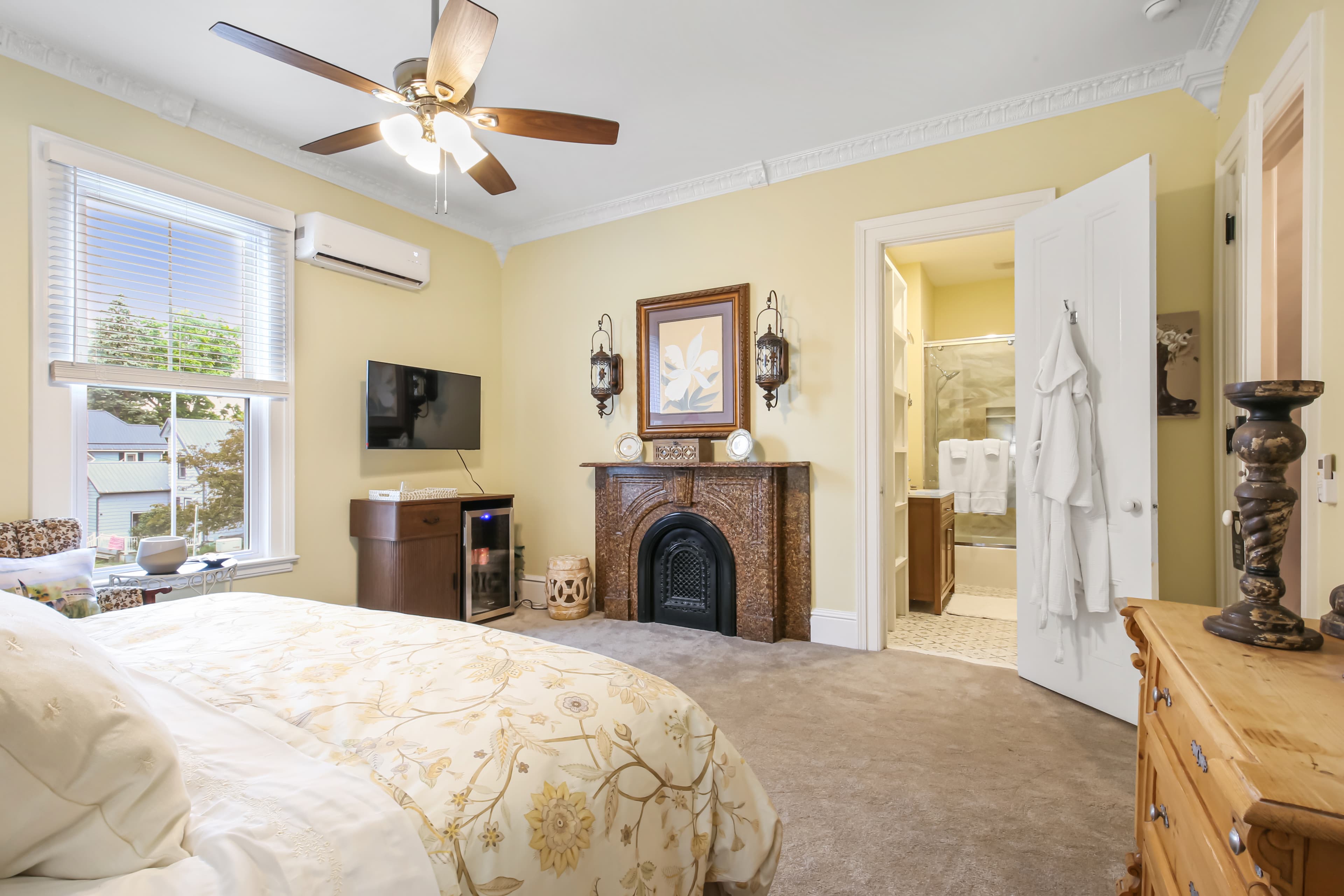 A cozy bedroom featuring a ceiling fan, decorative fireplace, and a view of an attached bathroom.