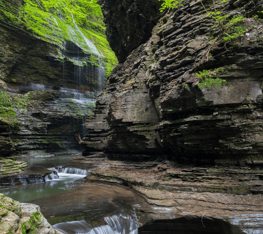 A serene waterfall cascades down rocky cliffs into a tranquil stream surrounded by lush greenery.